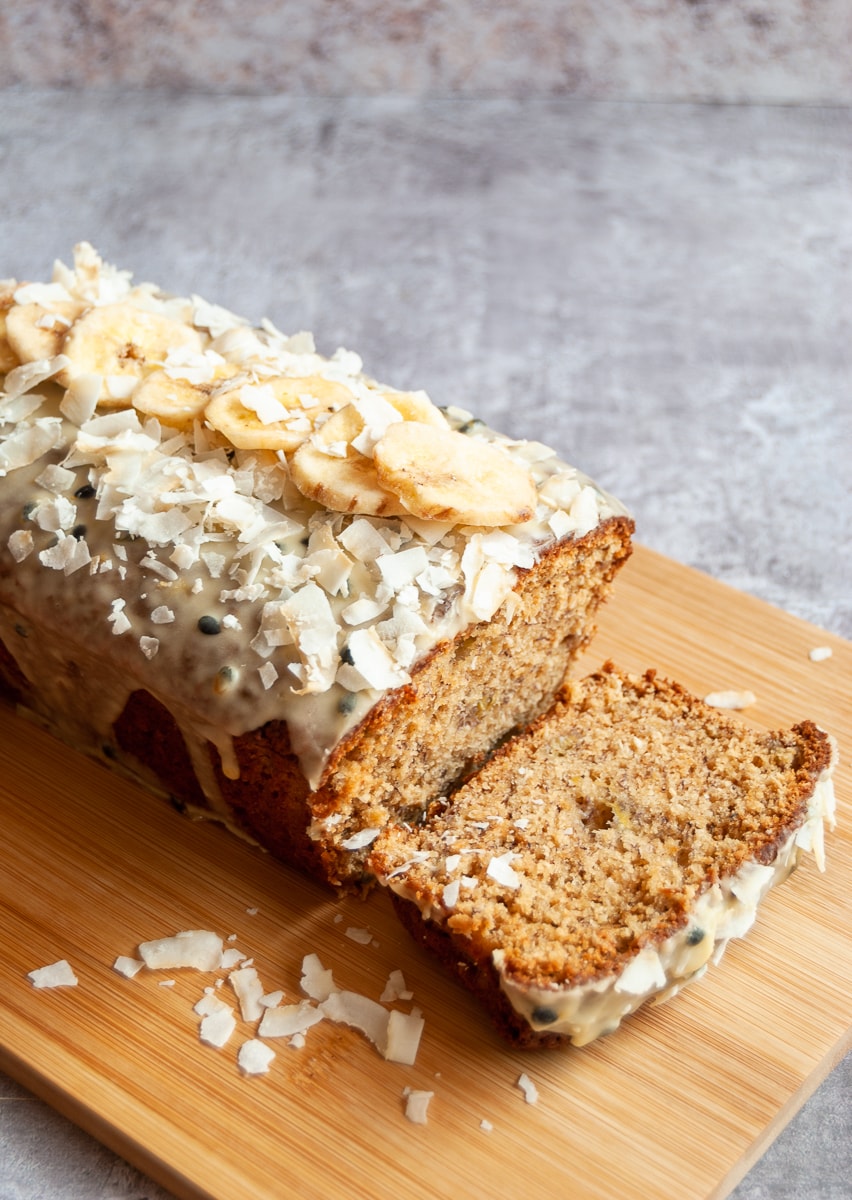 a banana and coconut cake topped with a passionfruit glaze, banana chips and coconut flakes on a wooden board