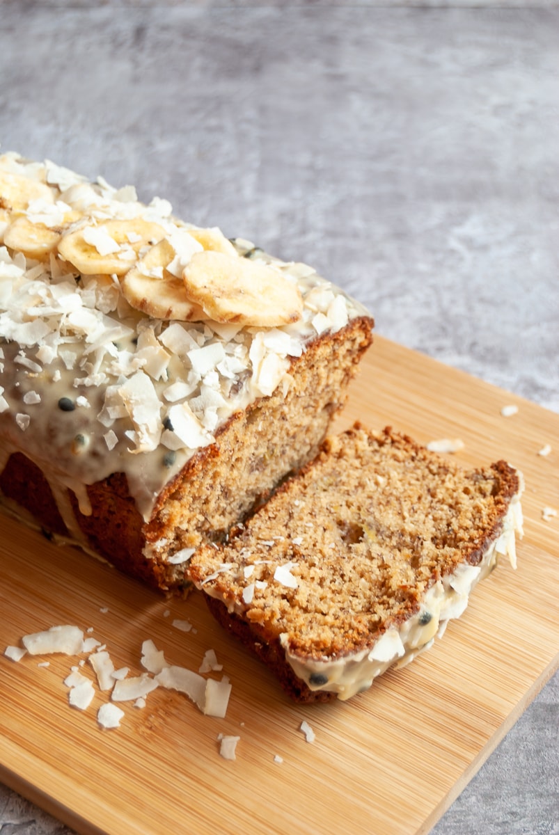 A banana and coconut cake covered in passionfruit glaze and dried banana chips and coconut flakes on a wooden board.