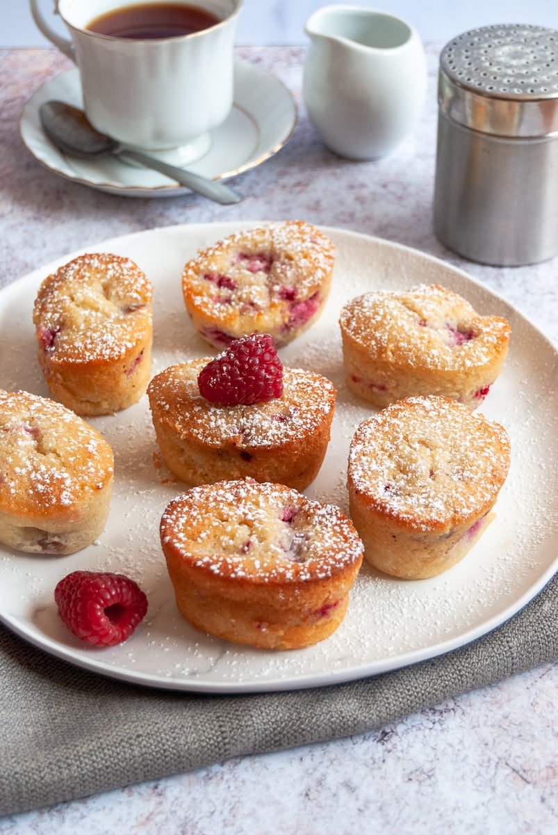 a white plate of raspberry and peach friands dusted with icing sugar, a cup of tea on a white saucer and a jug of milk