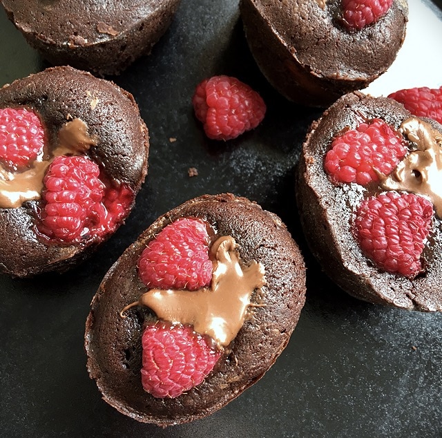A close up flat lay picture of gooey chocolate cakes with raspberries and chocolate hazelnut spread on a black plate.