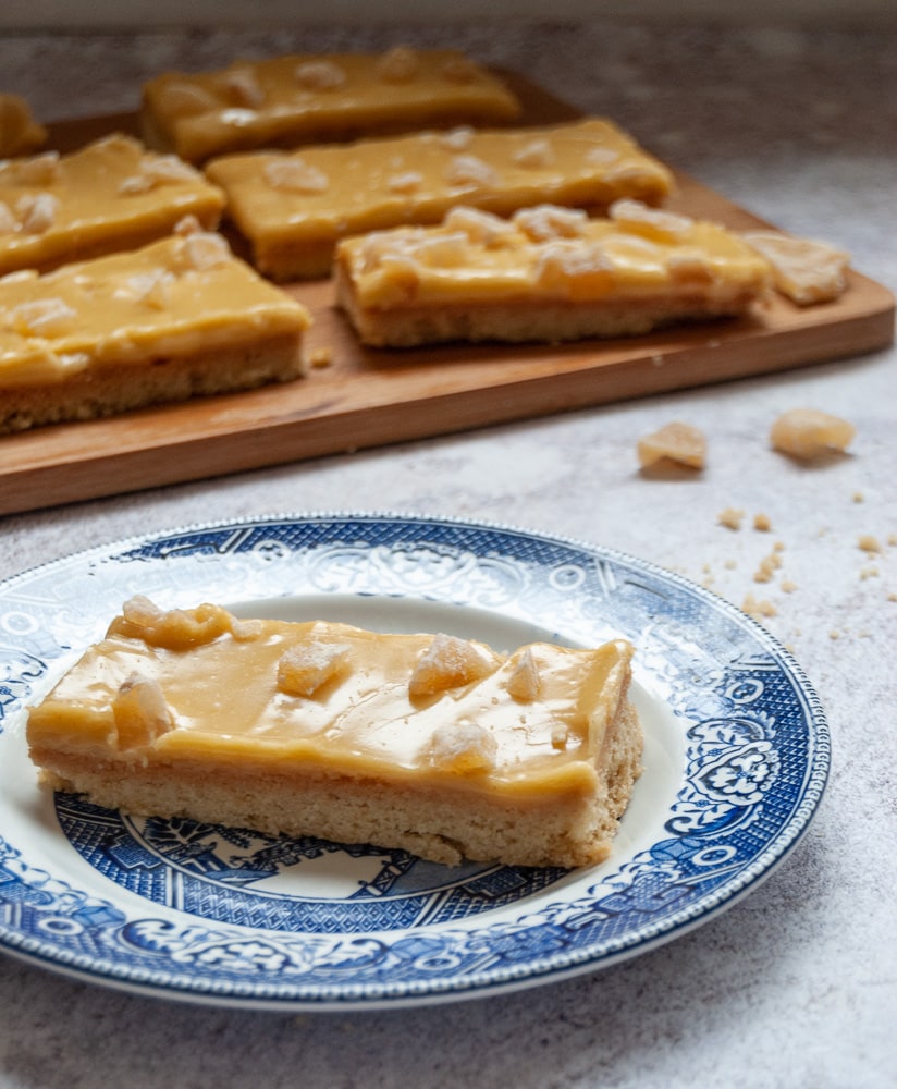 A slice of ginger crunch bar on a blue plate, with more bars on a wooden board in the background