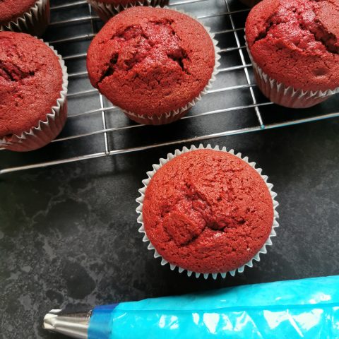 Red cupcakes on a wire rack with a blue piping bag of white frosting