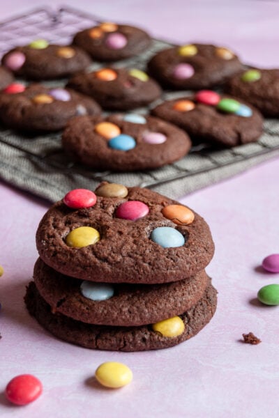 three chocolate cookies topped with smarties stacked on top of each other. A wire rack with more cookies can be seen in the background.