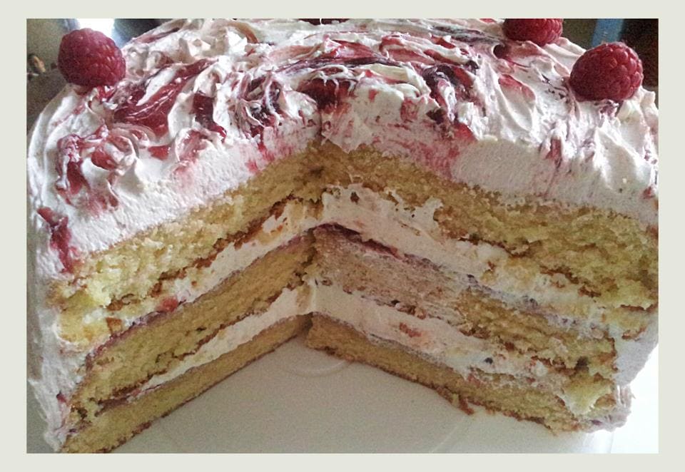 A three layer sponge cake sliced in half to reveal vanilla meringue icing and raspberry jam.