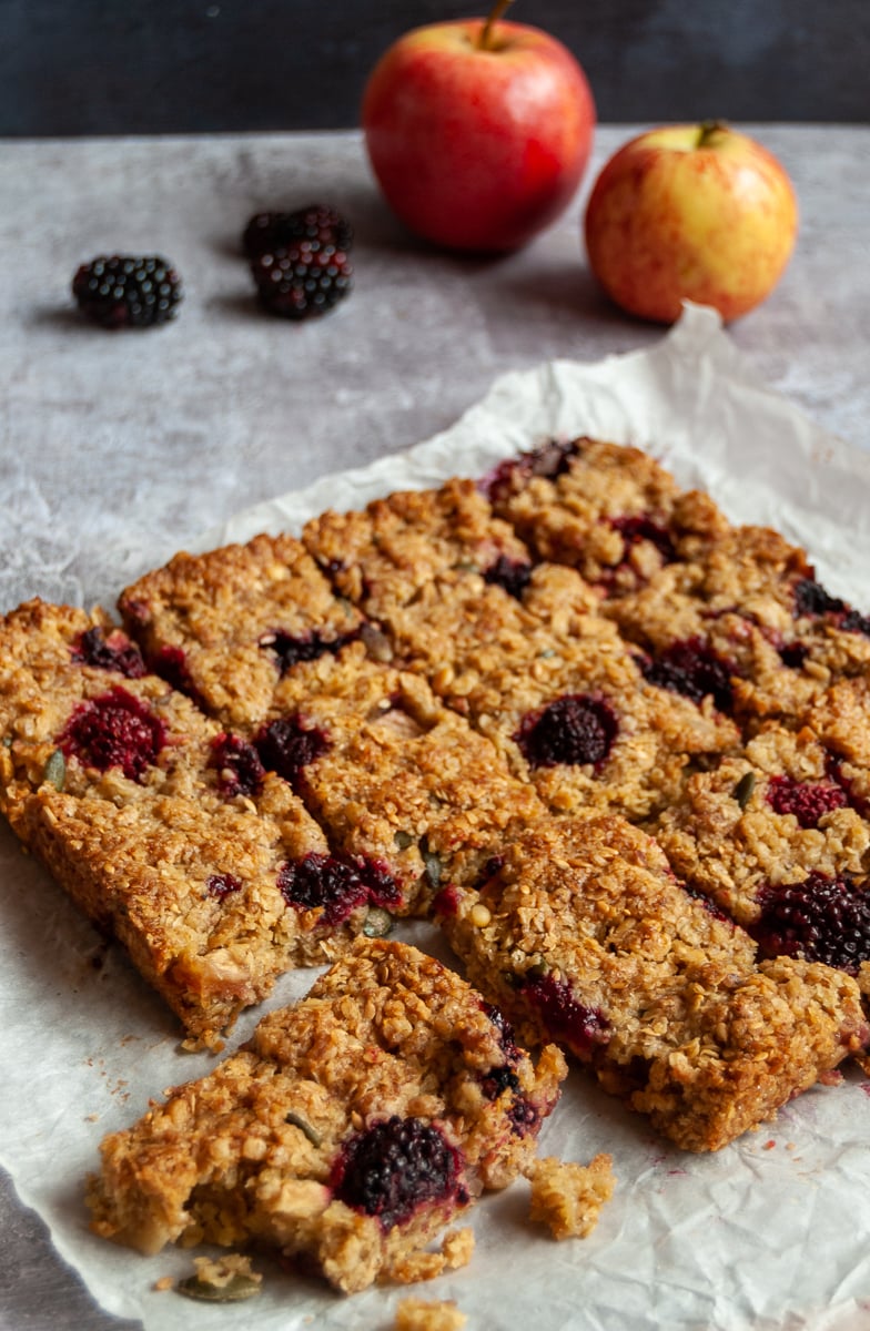 a batch of flapjack bars with apples and blackberries on a piece of baking parchment and two apples and blackberries in the background.