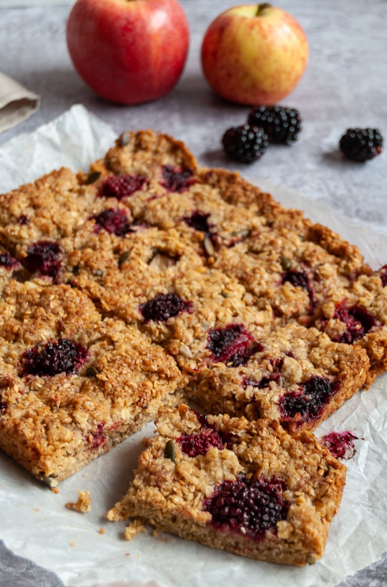apple and blackberry flapjack bars on a square of baking parchment with apples and blackberries in the background.