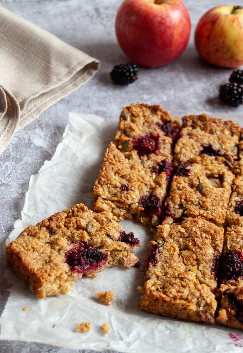 apple and blackberry studded oat bars on a piece of baking parchment, a cream linen napkin and two apples and fresh blackberries