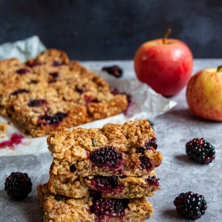 Three Flapjacks filled with apple and blackberries stacked on top of each other with more flapjacks on a sheet of baking parchement in the background. Two apples and blackberries are sitting beside the flapjacks.