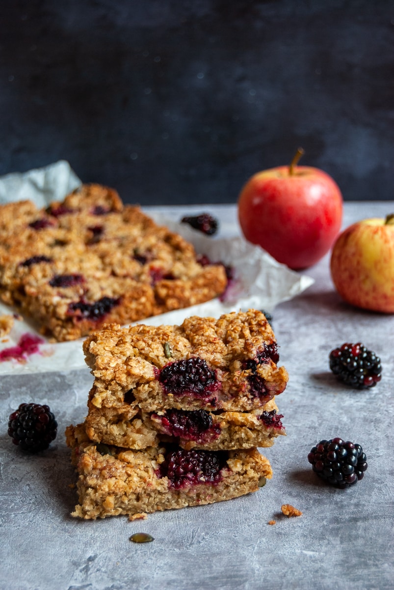 Three Flapjacks filled with apple and blackberries stacked on top of each other with more flapjacks on a sheet of baking parchement in the background. Two apples and blackberries are sitting beside the flapjacks.