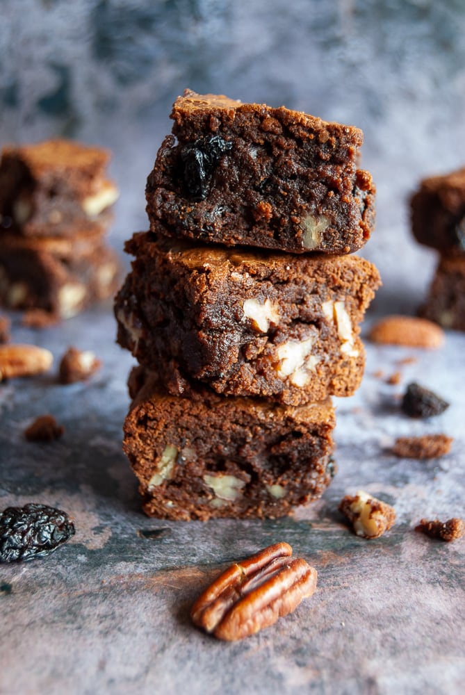 Three cherry and pecan brownies piled on top of eachother. Pecan nuts and dried cherries are scattered around the brownies, background is a mottled grey.