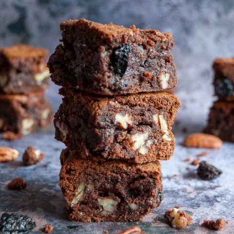 Three cherry and pecan brownies piled on top of eachother. Pecan nuts and dried cherries are scattered around the brownies, background is a mottled grey.