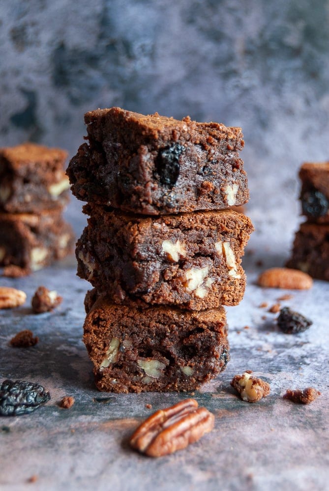 Three cherry and pecan brownies piled on top of eachother. Pecan nuts and dried cherries are scattered around the brownies, background is a mottled grey.