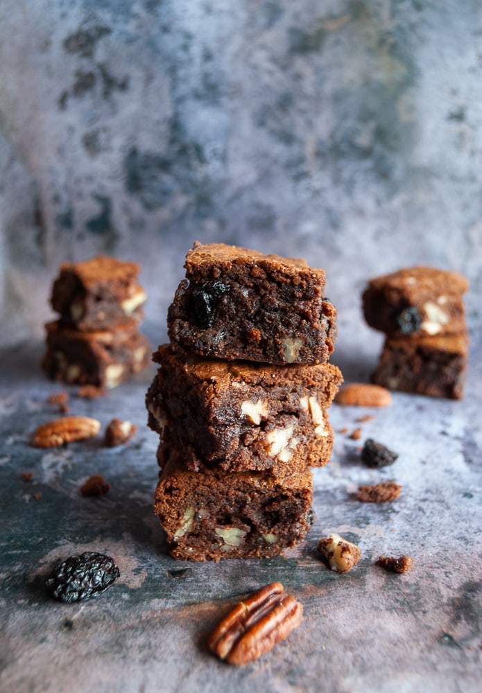 Three cherry and pecan brownies piled on top of eachother. Pecan nuts and dried cherries are scattered around the brownies. More brownies can be seen in the background. 