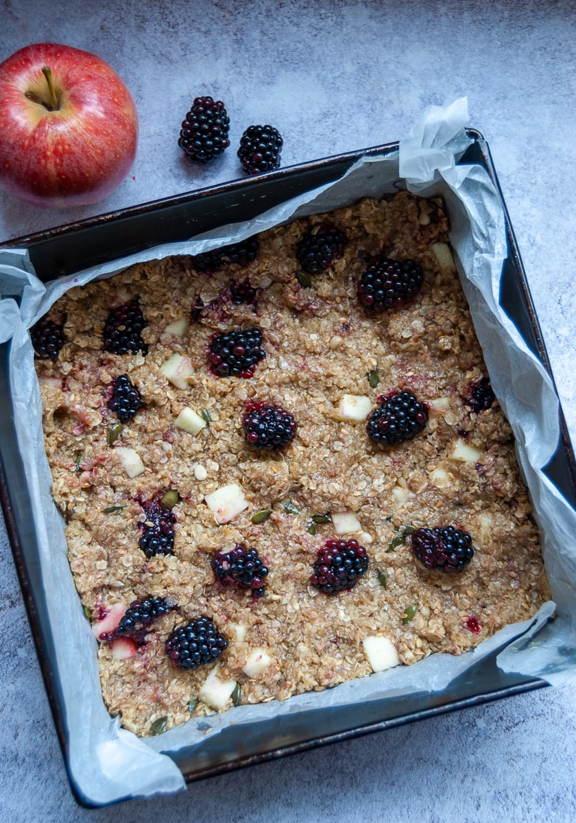 A tin of unbaked apple and blackberry flapjacks, a red apple and a couple of blackberries