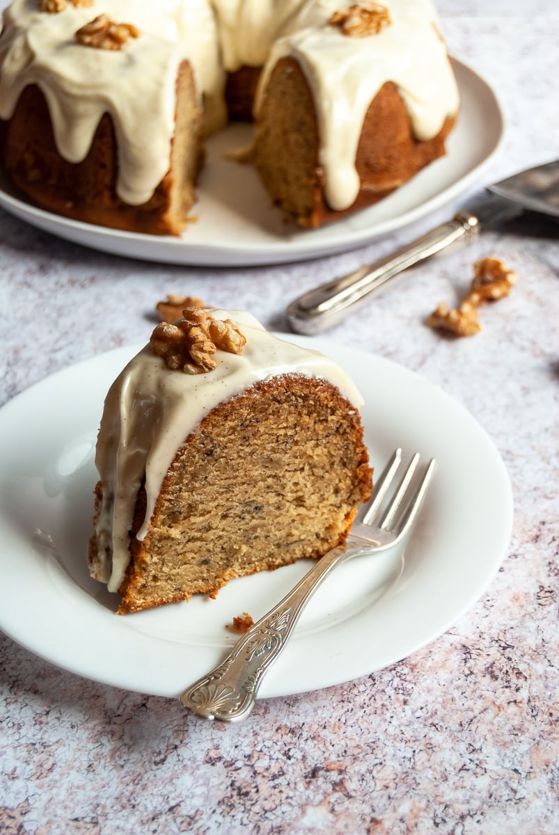 a slice of banana bundt cake topped with cream cheese glaze and a walnut on a white plate with a cake fork. A large banana bundt cake in the background.