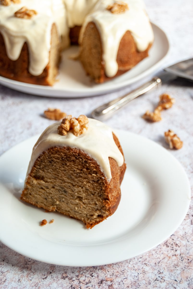 A slice of banana cake topped with cream cheese glaze and a walnut on a white plate and a banana bundt cake in the background.