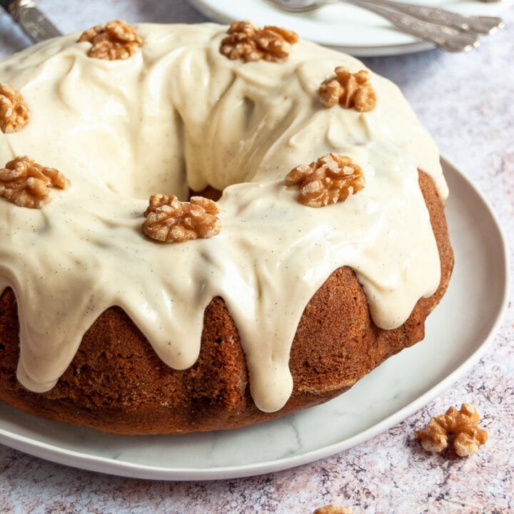 A banana bundt cake topped with vanilla cream cheese glaze and walnuts on a white plate