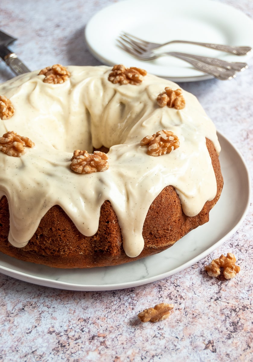 A banana bundt cake topped with vanilla cream cheese glaze and walnuts on a white plate