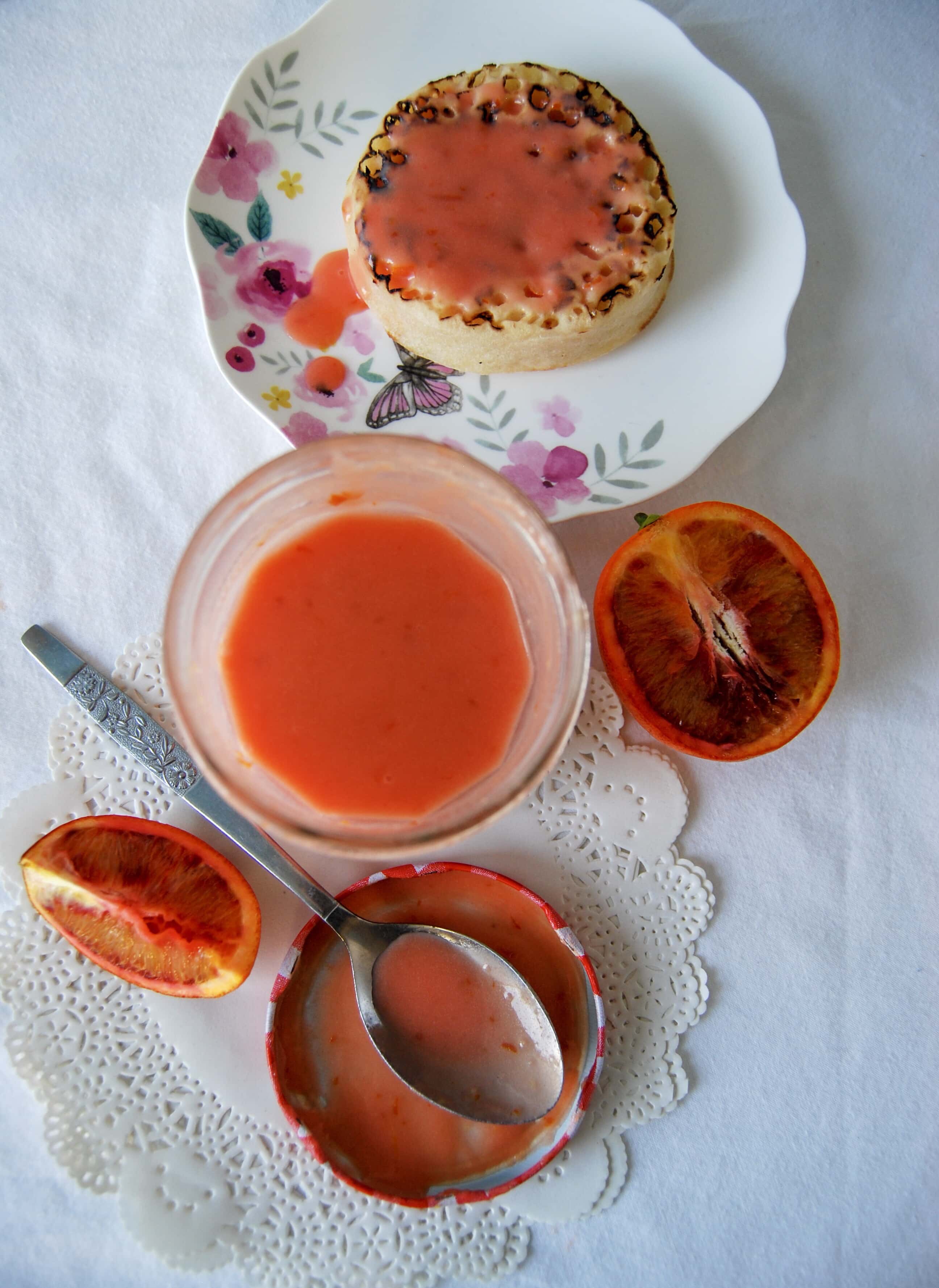 A flat lay picture of a jar of blood orange curd with a crumpet spread with blood orange curd on a white tablecloth.