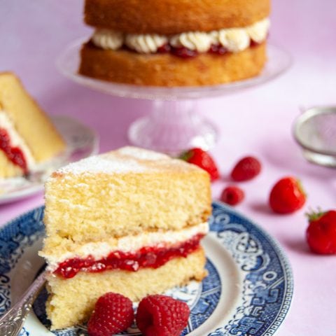 A photo of a slice of Victoria sponge with a raspberry jam and fresh cream filling on a blue willow pattern plate. The victoria sponge cake can also be seen in the background on a glass cake stand. Fresh berries are scattered around for a decorative effect.