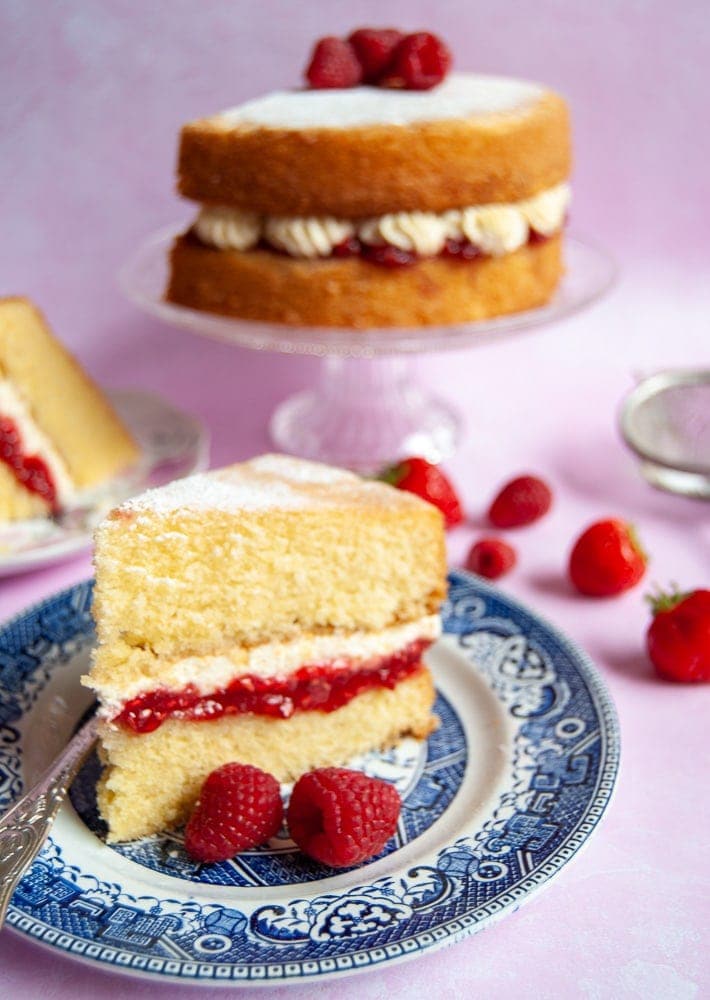 A photo of a slice of Victoria sponge with a raspberry jam and fresh cream filling on a blue willow pattern plate. The victoria sponge cake can also be seen in the background on a glass cake stand. Fresh berries are scattered around for a decorative effect.