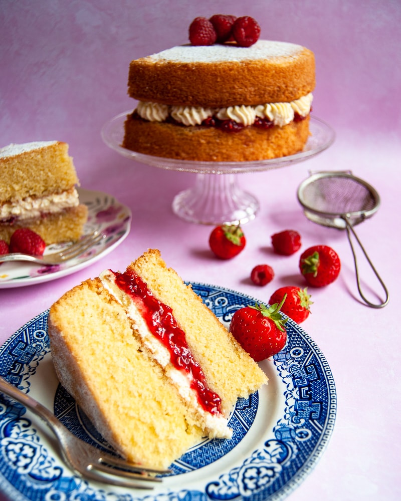 A Photo of a cut slice of Victoria Sponge Cake filled with raspberry jam and whipped cream on a blue willow pattern plate. The rest of the cake on a glass cake stand can be seen in the background.