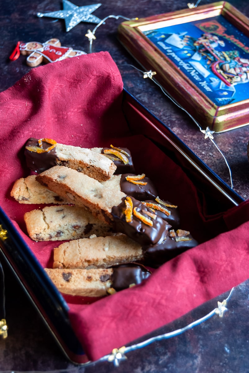 a box lined with a red napkin of biscotti cookies coated in chocolate and sprinkled with candied orange peel.  A silver star and a gingerbread lady can be seen in the background