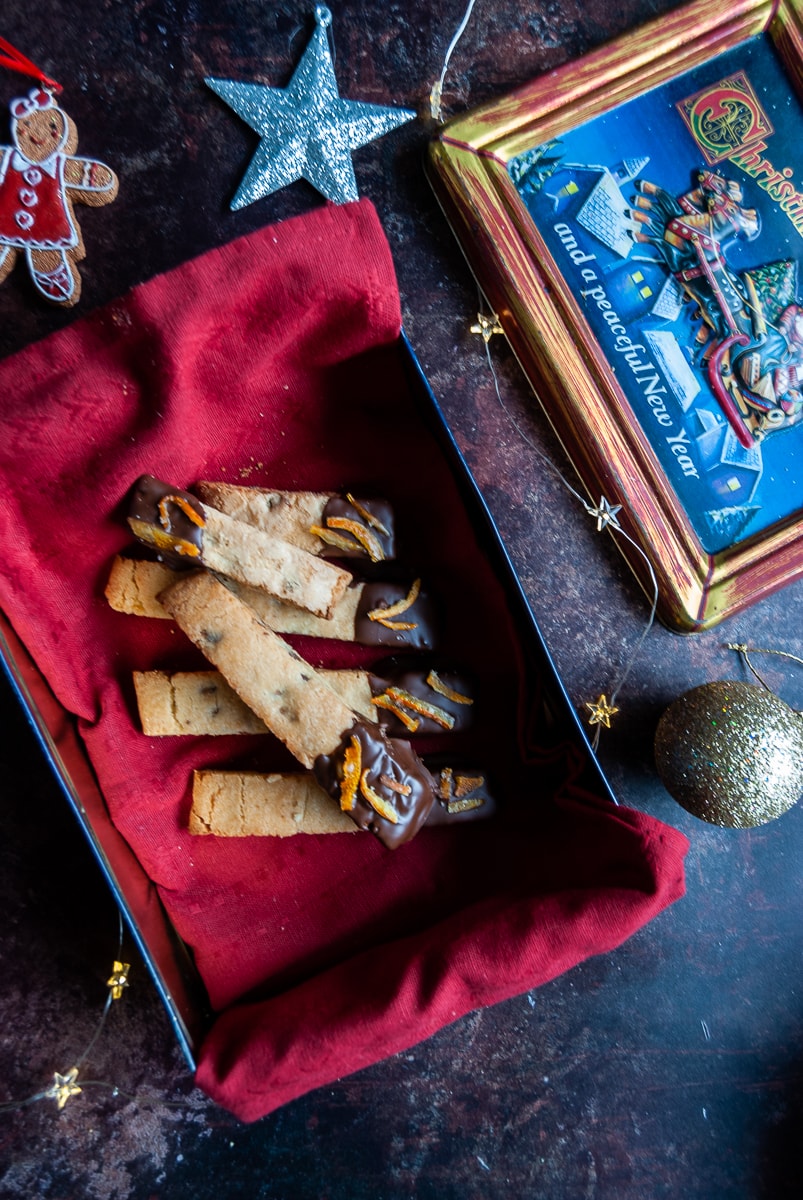 a flat lay photo of a box lined with a red napkin with chocolate dipped biscotti cookies inside. Christmas decorations are placed around the box for a festive scene.