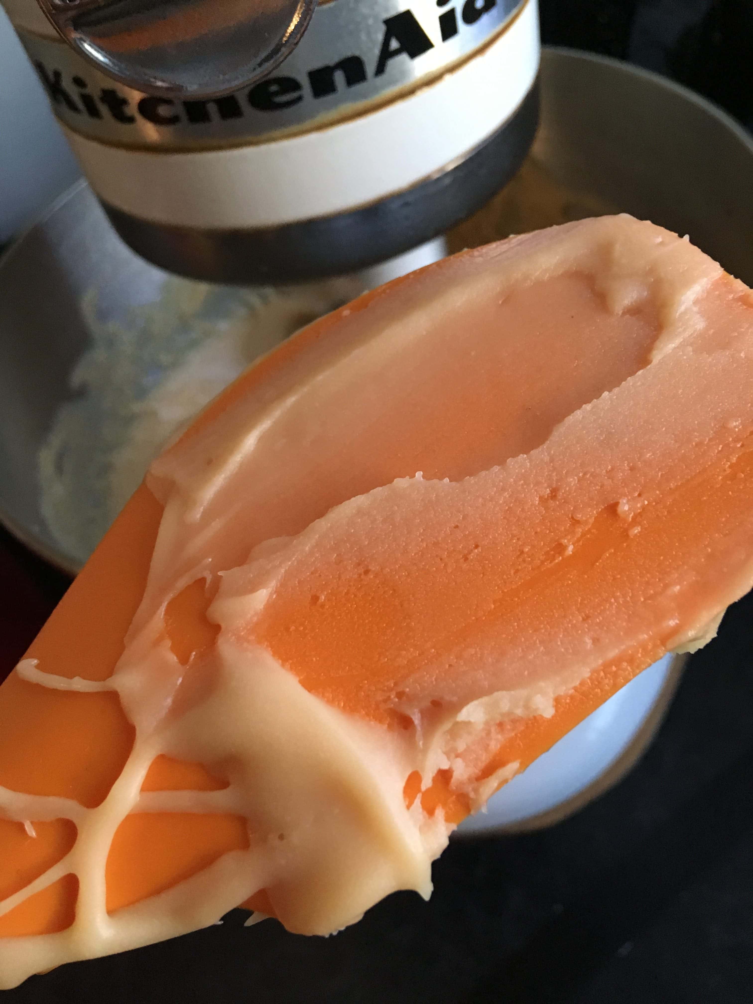 An orange spatula covered in Scottish tablet mixture to show the desired consistency and a Kitchen Aid stand mixer.