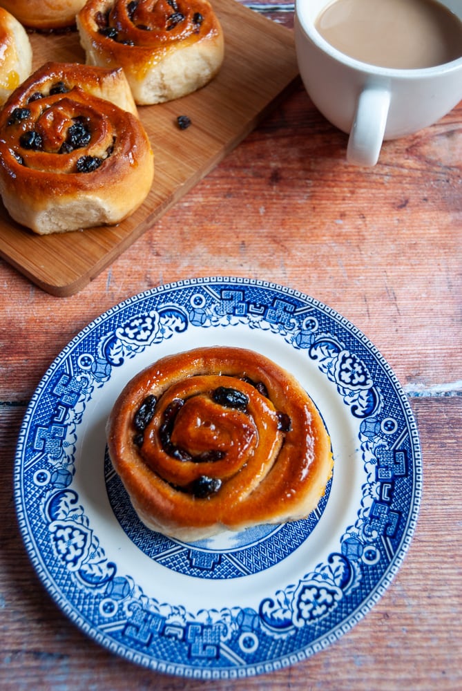 A flat lay photo of a Chelsea bun on a blue and white Plate, a cup of coffee and more buns on a wooden board 