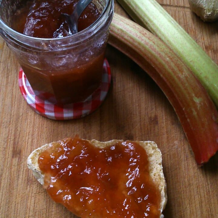 a jar of rhubarb jam with a spoon resting in the jar, two sticks of rhubarb and a scone spread with the jam on a wooden board.