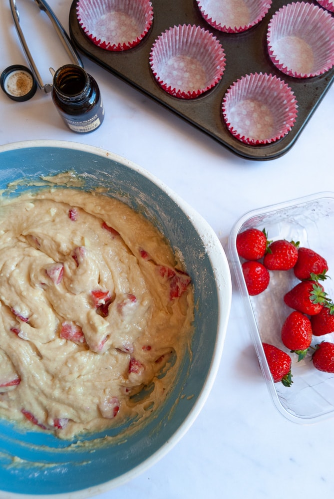 a blue and white bowl of strawberry muffin batter, a container of fresh strawberries, a muffin pan lined with red muffin liners, an ice cream scoop and a bottle of vanilla bean paste on a white backdrop.