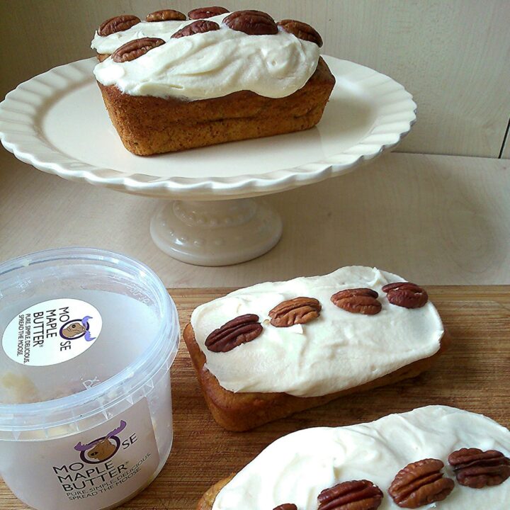 three small banana loaf cakes topped with cream cheese icing and pecan nuts on a wooden board and cream cake stand