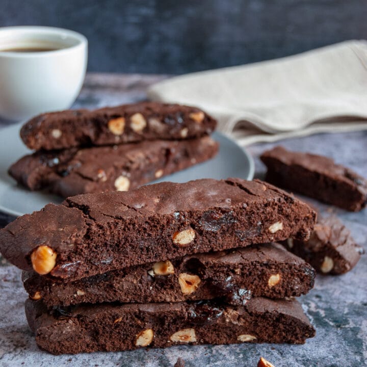 three chocolate hazelnut biscotti stacked on top of each other on a grey backdrop. A white cup of coffee and a beige linen napkin in the background of the picture.