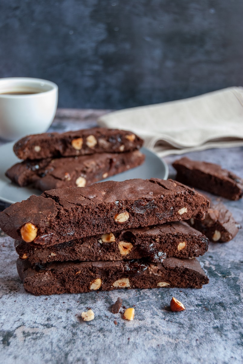 three chocolate hazelnut biscotti stacked on top of each other on a grey backdrop. A white cup of coffee and a beige linen napkin in the background of the picture.