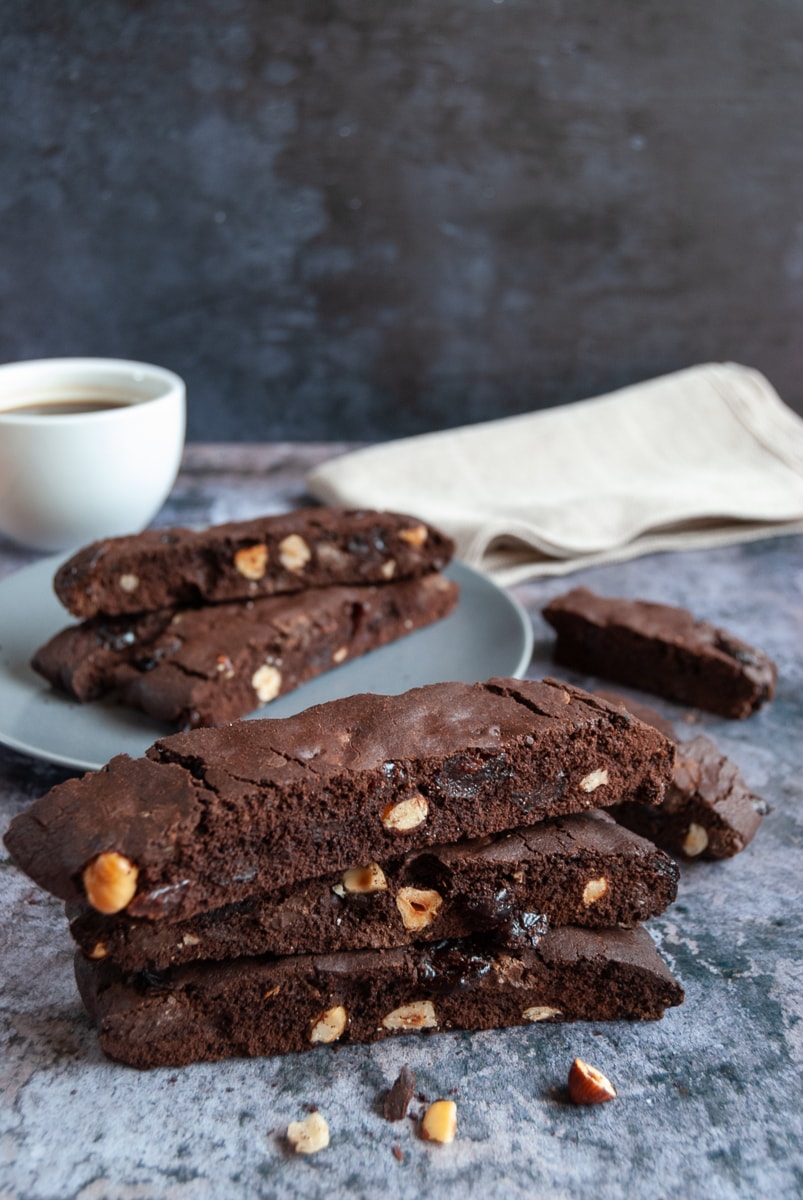 a stack of three chocolate and hazelnut biscotti on a wooden board, a white cup of coffee and a saucer with a piece of broken biscotti and a beige linen napkin