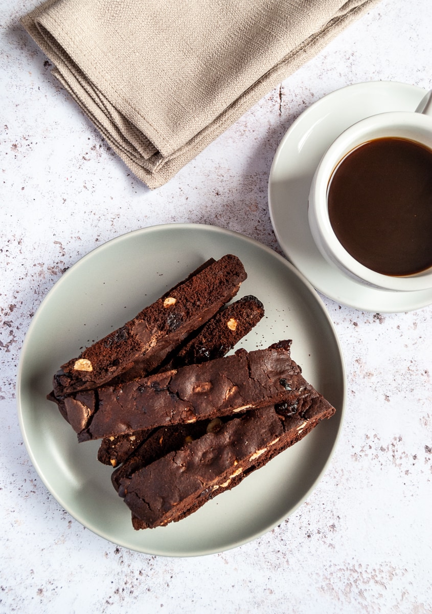 A flat lay photo of a grey plate of chocolate biscotti, a white cup of coffee on a white saucer with a piece of biscotti and a beige linen napkin