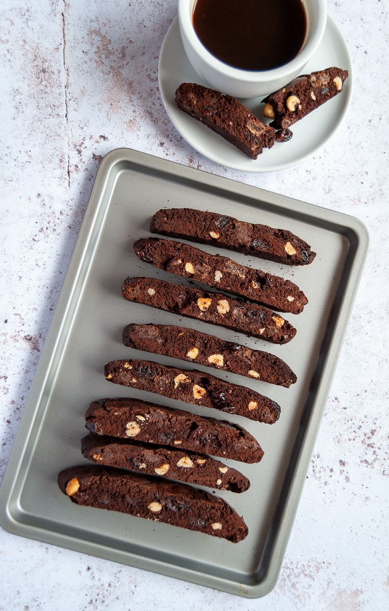 A flat lay photo of a silver tray of chocolate and hazelnut biscotti and a white cup of coffee with a saucer and a piece of biscotti