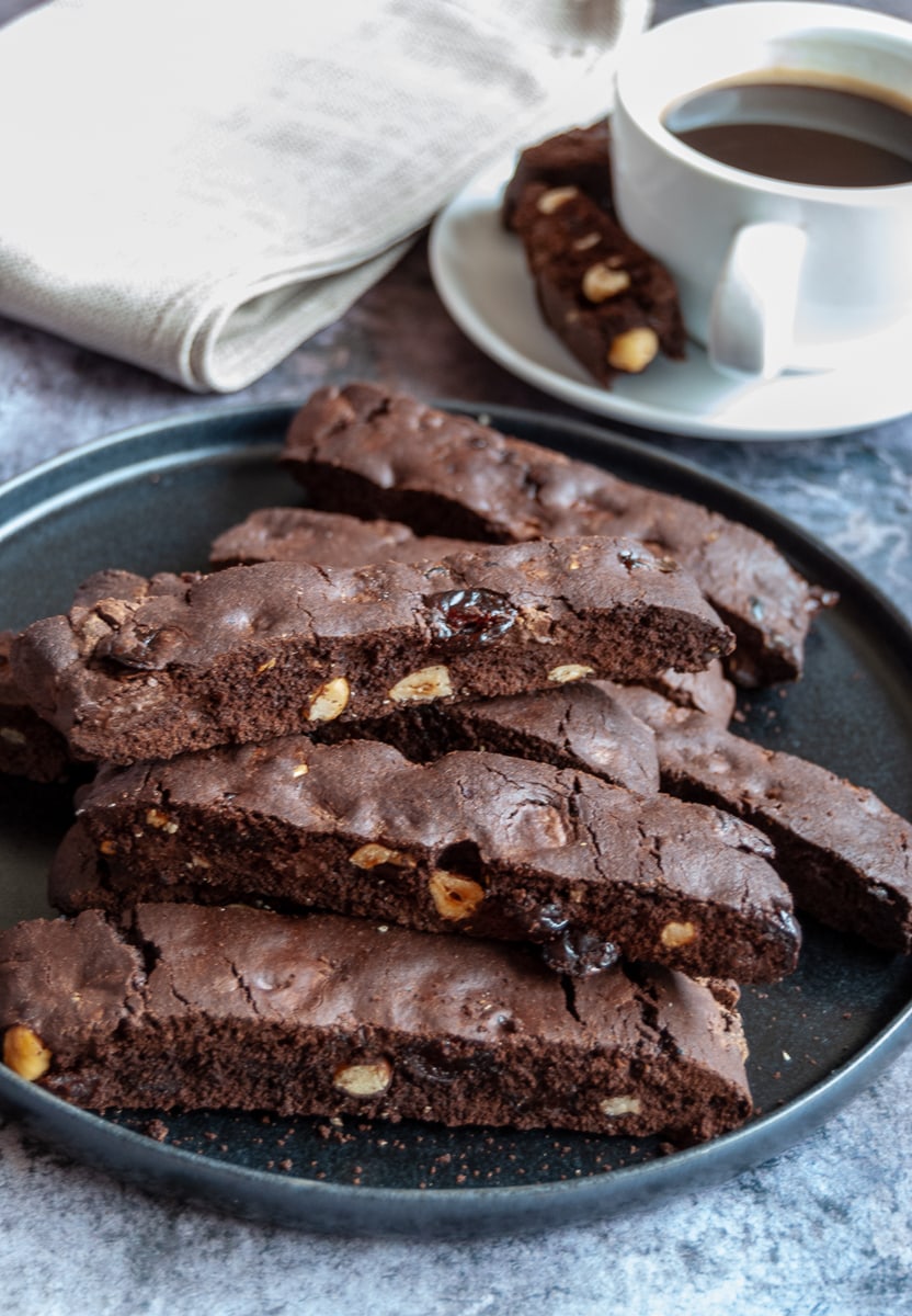 A black plate of chocolate and hazelnut biscotti, a white cup of coffee and saucer with more biscotti and a beige linen napkin.