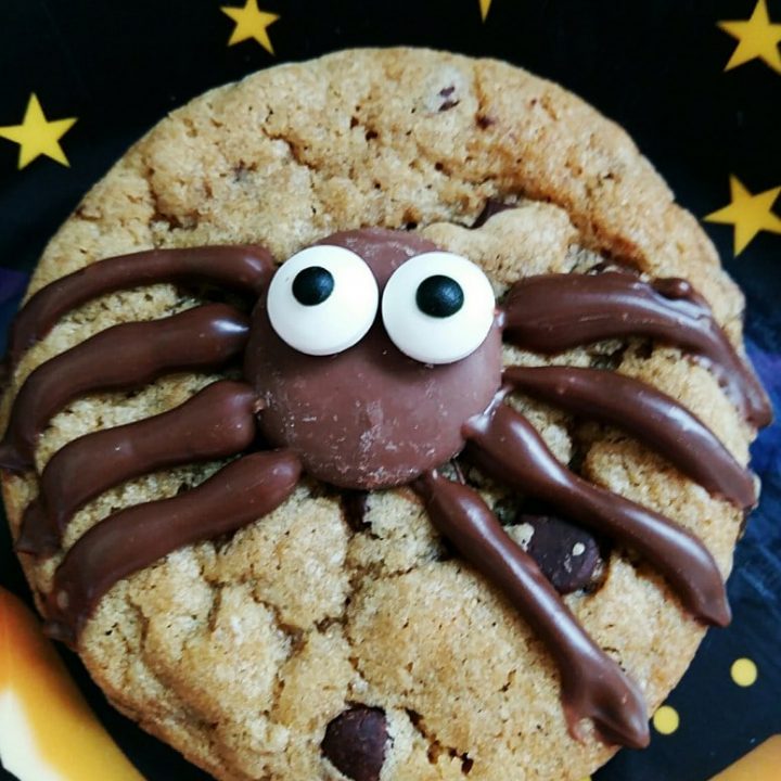 a close up image of a chocolate chip cookie topped with a spider made on melted chocolate, a chocolate button and candy eyes.