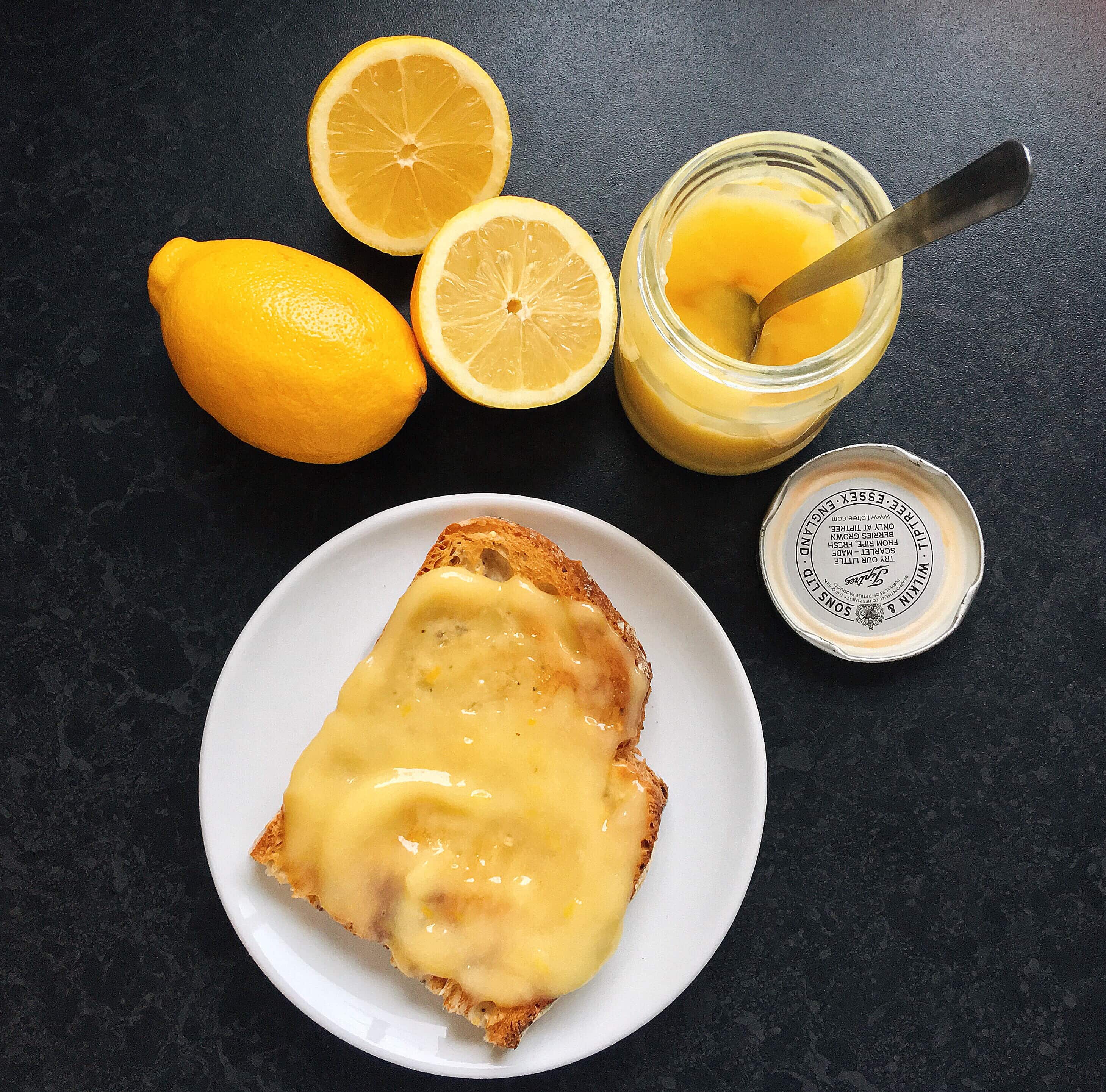 A slice of toast on a white plate spread with lemon curd, a jar of lemon curd with a spoon and two lemons, one cut in half.