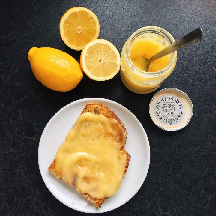 A jar of lemon curd with a spoon and a slice of toast spread with lemon curd