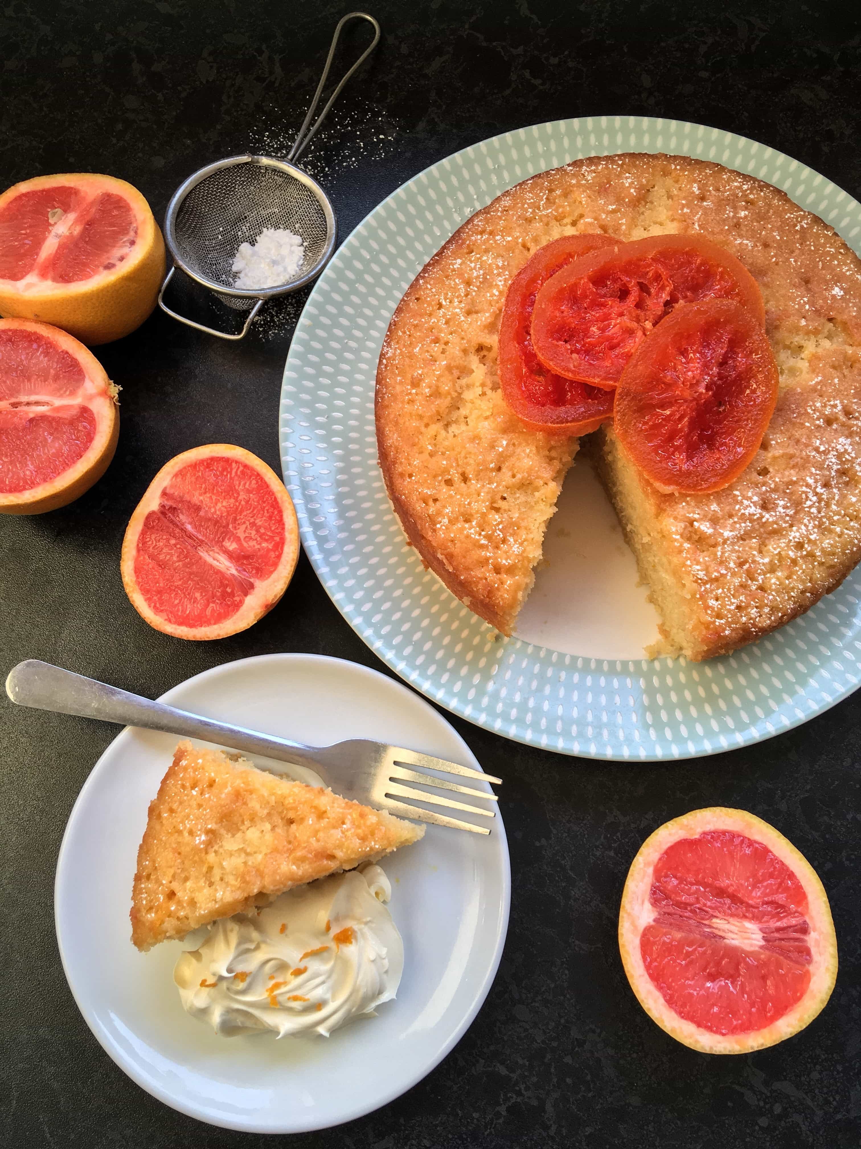 A pink grapefruit drizzle cake decorated with candied grapefruit slices. A slice of the cake with whipped cream and halved grapefruits.