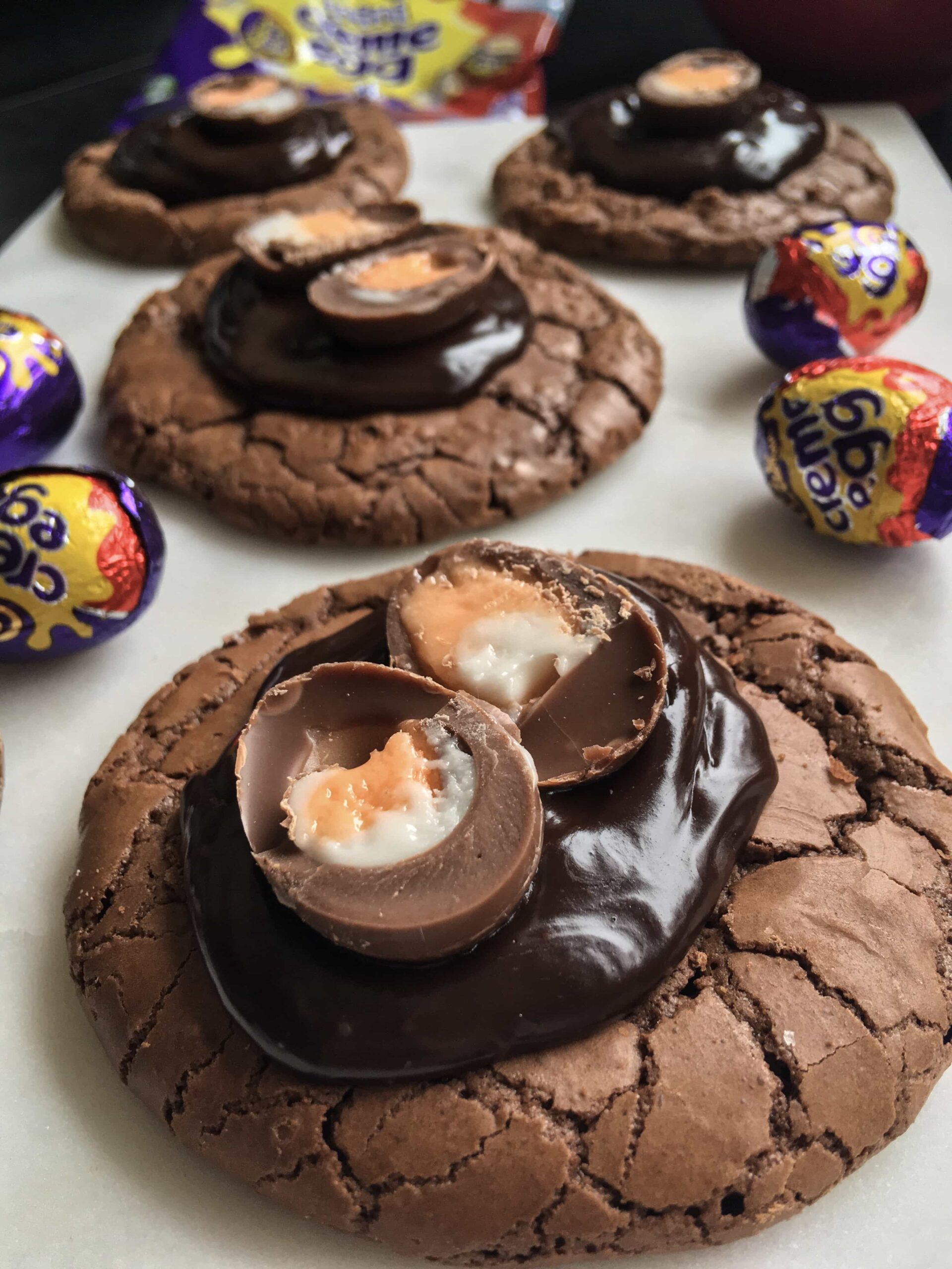 a close up picture of chocolate brownie cookies topped with chocolate ganache and a mini creme egg on a white board