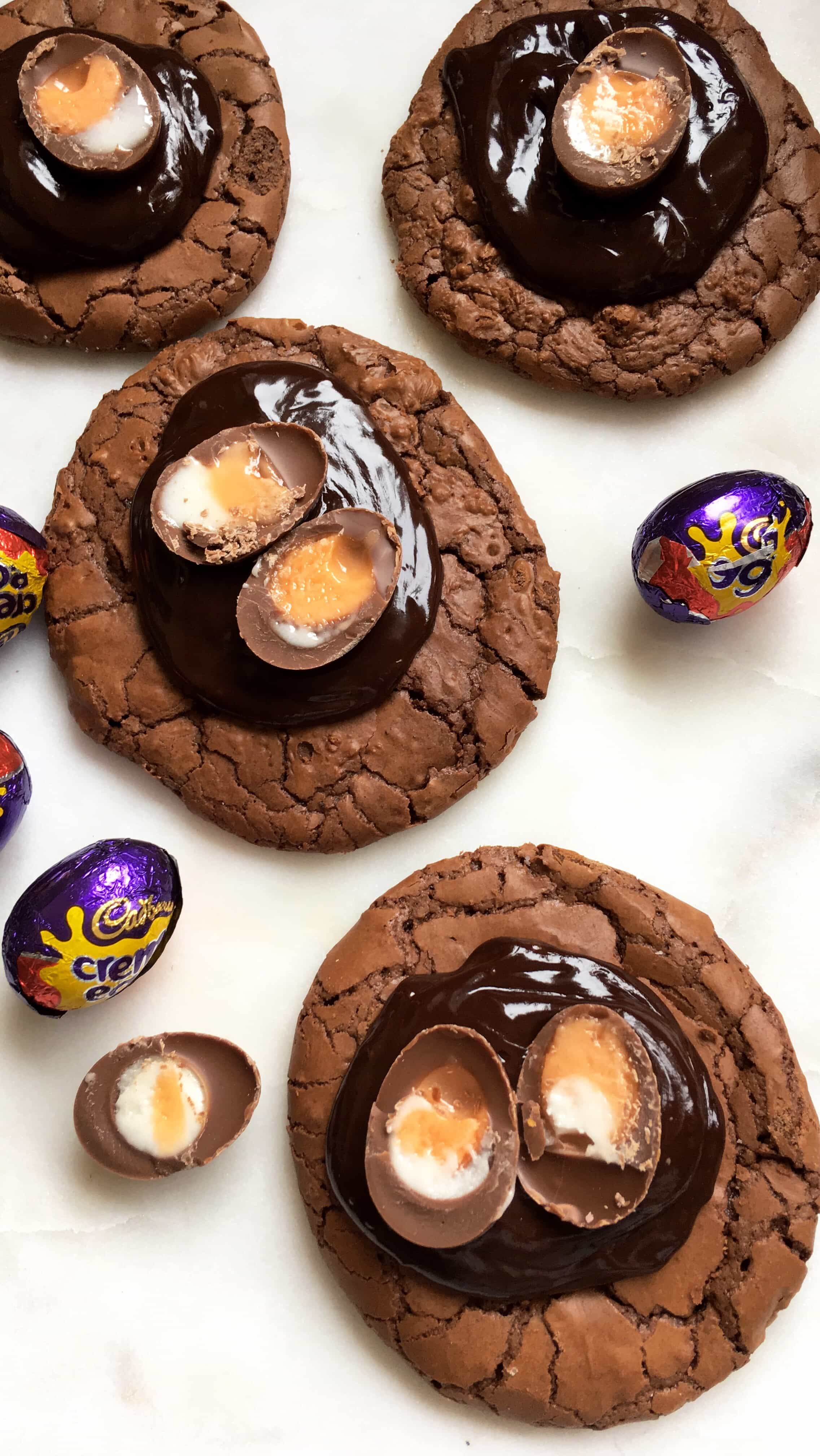 four chocolate brownie cookies topped with chocolate ganache and a mini creme egg on a white board