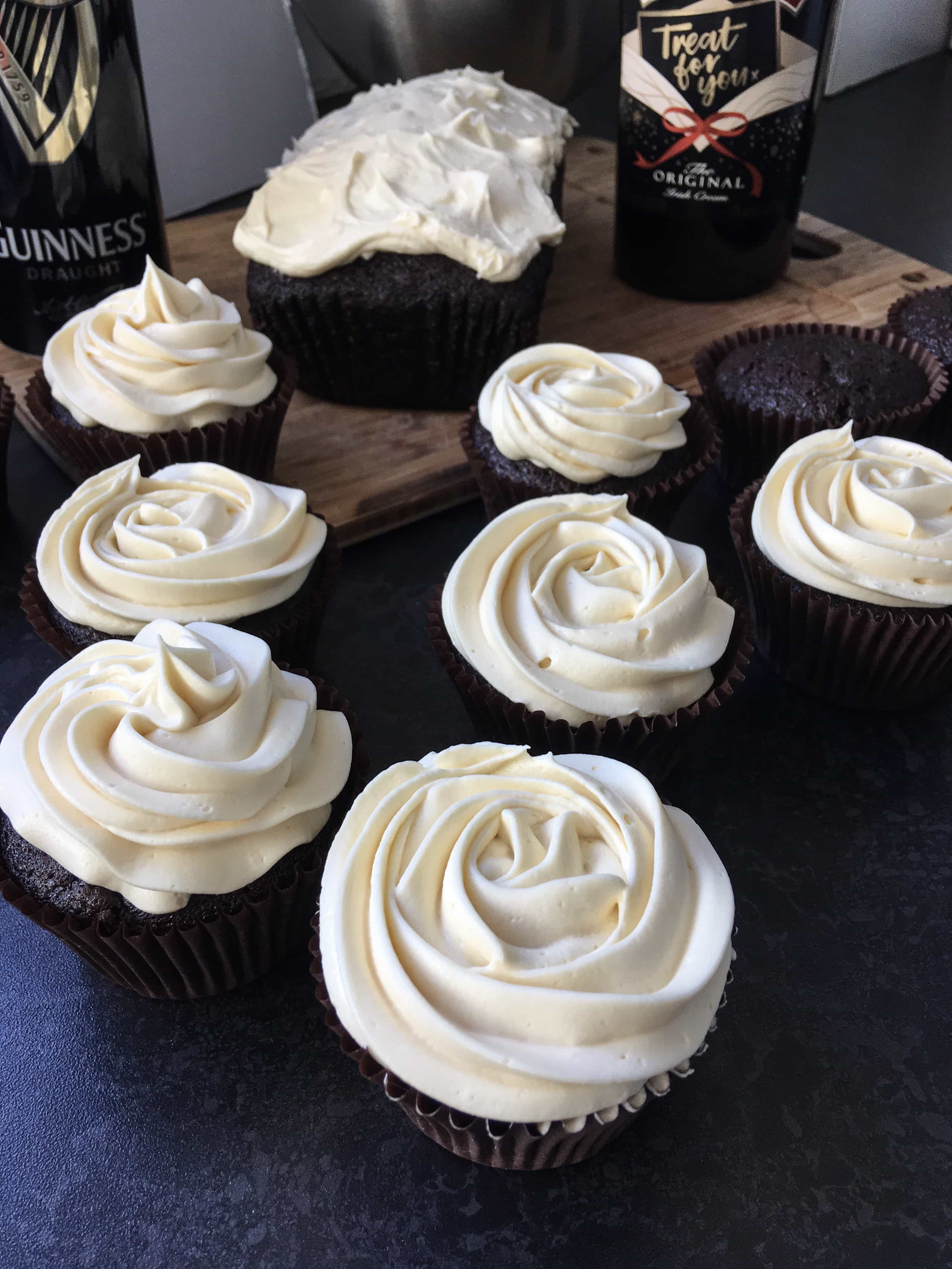 a row of chocolate cupcakes topped with a rose swirl of baileys frosting. A chocolate loaf cake topped with the frosting on a wooden board with a can of Guinness and a bottle of Baileys Irish cream