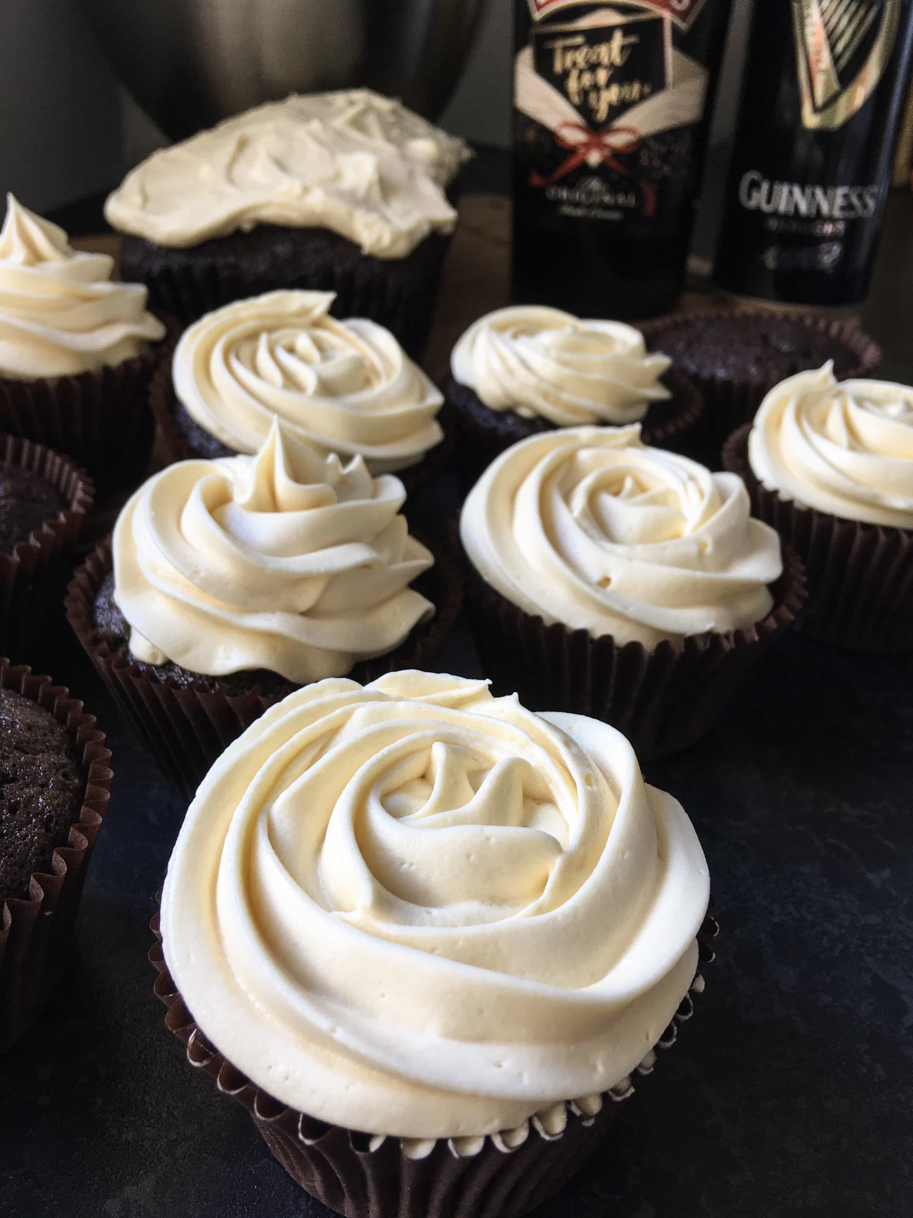 A close up photo of dark chocolate cupcakes topped with a rose style swirl of Baileys cream frosting. A bottle of Baileys and a Can of Guinness can be seen in the background