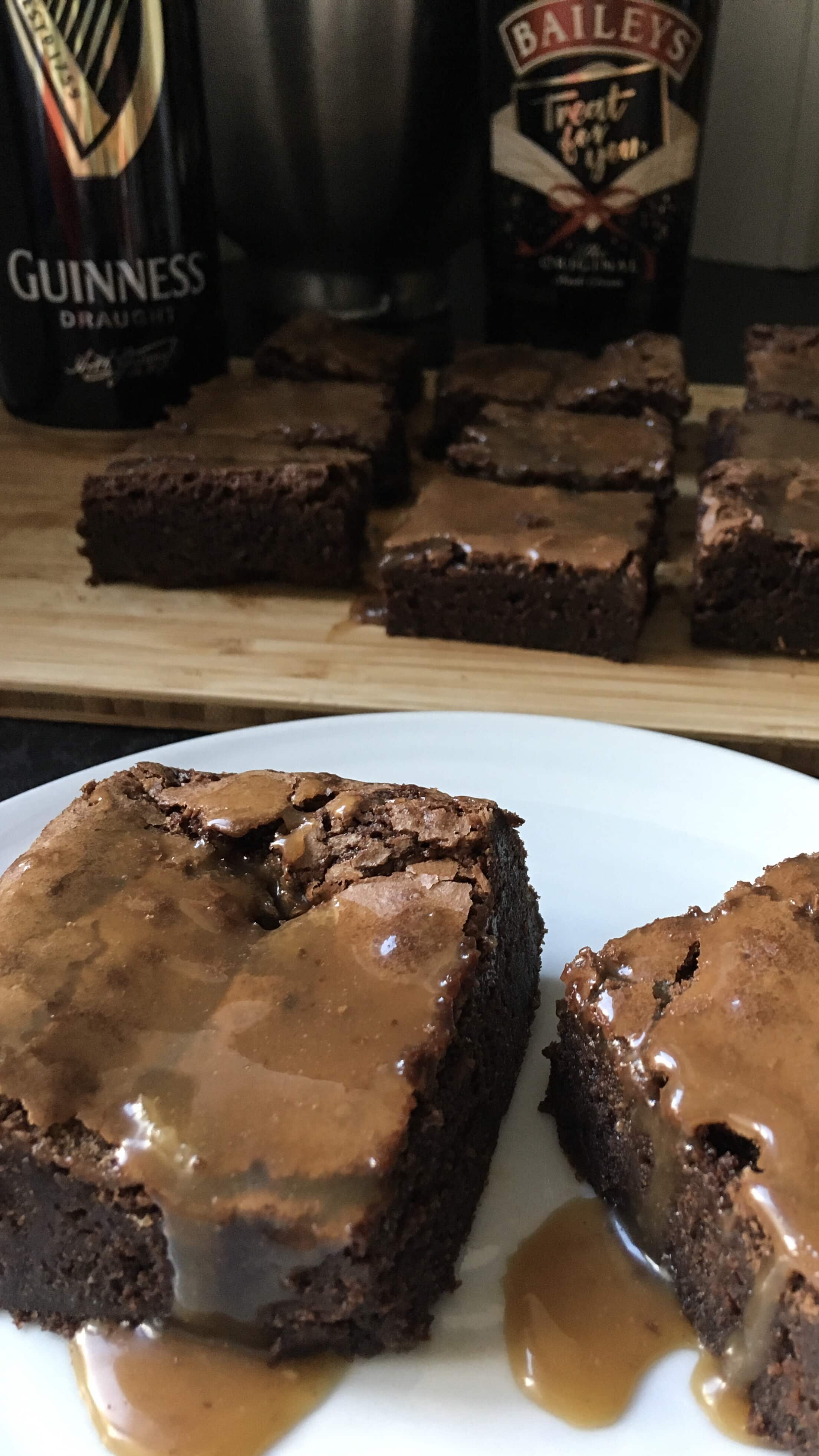 Two Guinness brownies with a salted caramel Bailey's Glaze on a white plate. More brownies and a bottle of Bailey's Irish cream and a can of Guinness can be seen in the background.