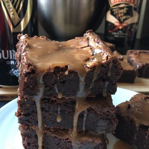 three Guinness brownies covered in a salted caramel glaze on a white plate. A can of Guinness and a bottle of Baileys can be seen in the background.