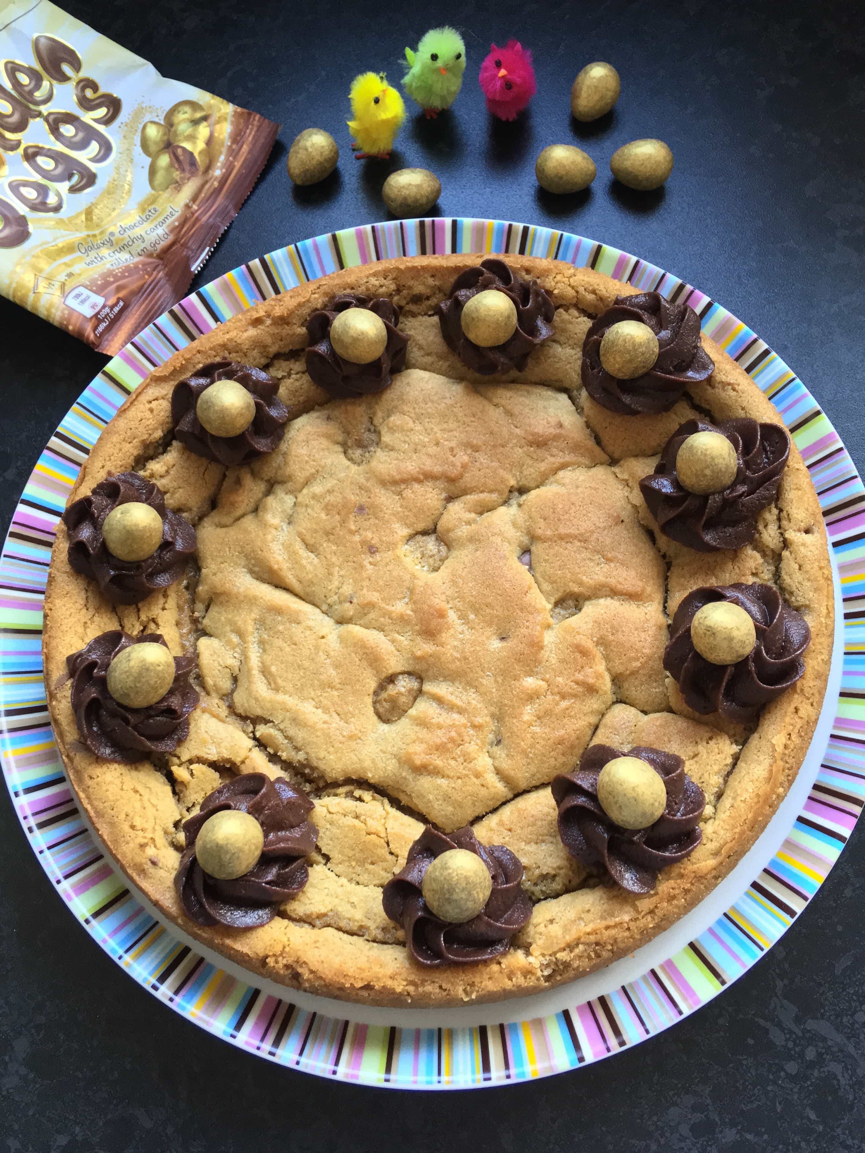 a giant chocolate chunk cookie cake with chocolate buttercream swirls and golden chocolate eggs on a striped plate